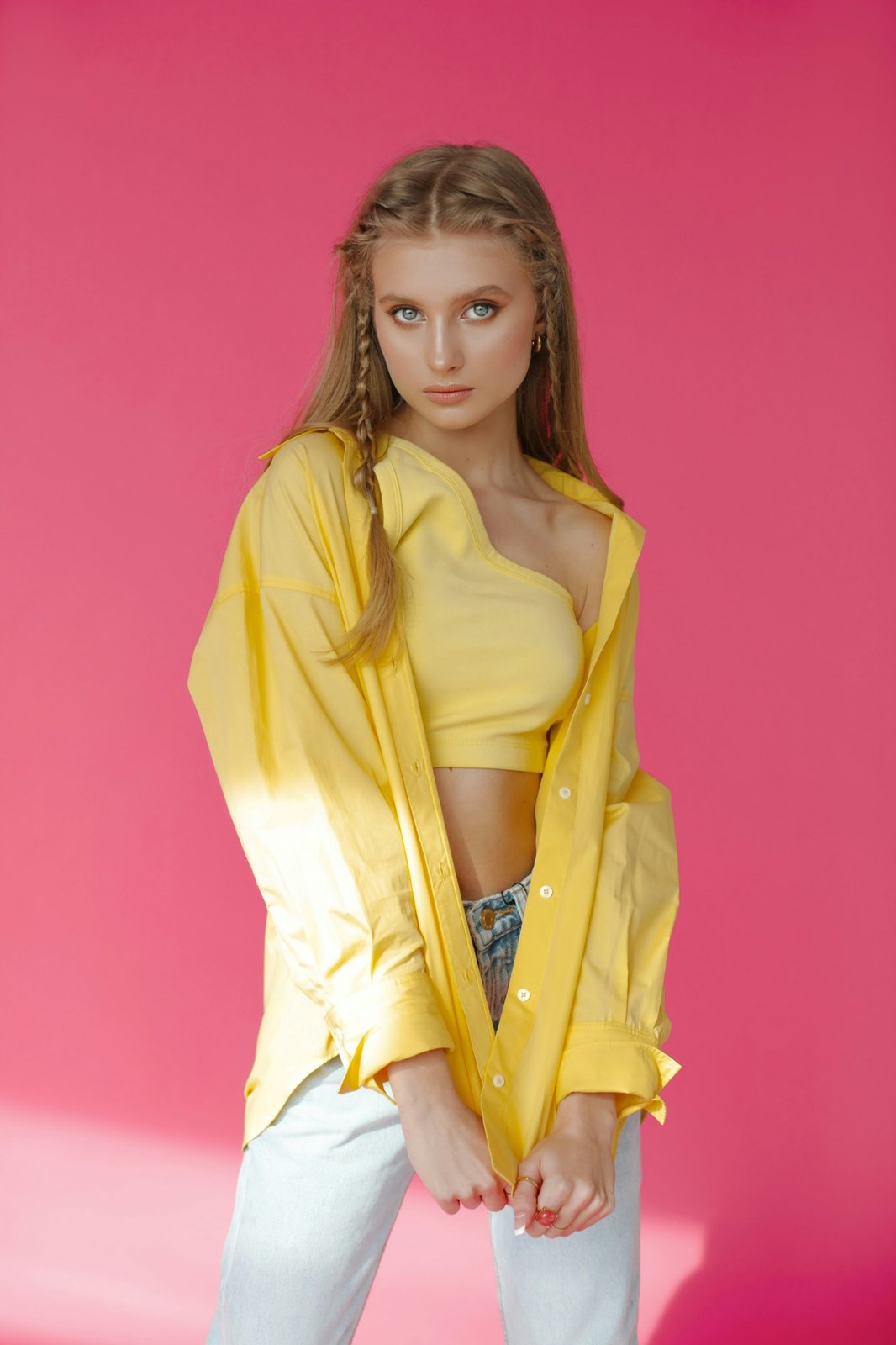 A young woman with long blonde hair and two thin braids, in an oversized yellow shirt, in front of a saturated pink studio backdrop